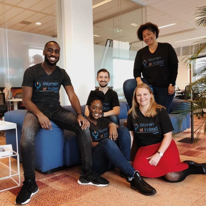 Image of five Indeed inclusion group members smiling and wearing shirts labeled "Women at Indeed" in a relaxed office setting